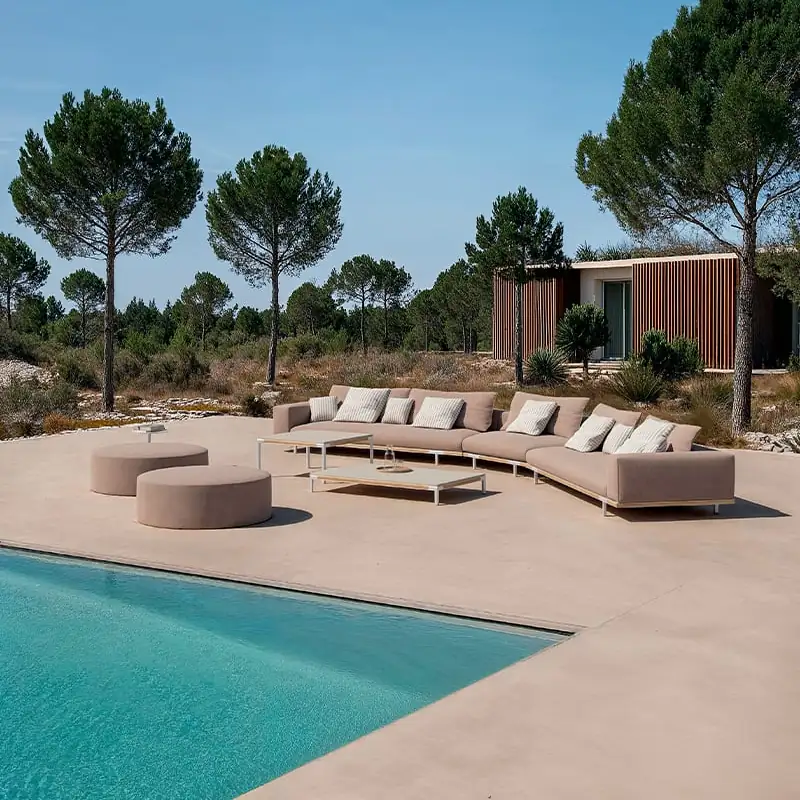 Ethimo Costiera long curved sofa on a sandy terrace beside a turquoise pool, with umbrella pines against a blue sky