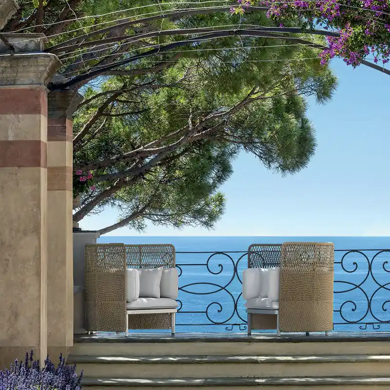 Two Ethimo Esedra high-back woven armchairs on a tiled balcony with an iron railing, bougainvillea and a pine above the sea