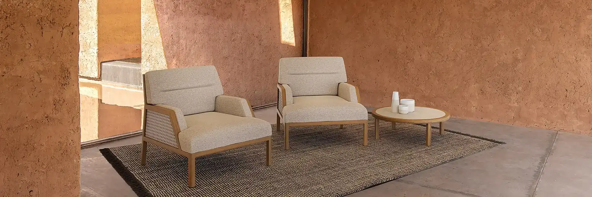 Two Ethimo Grand Life armchairs with pale cushions and a side table on a rug in a terracotta-walled loggia