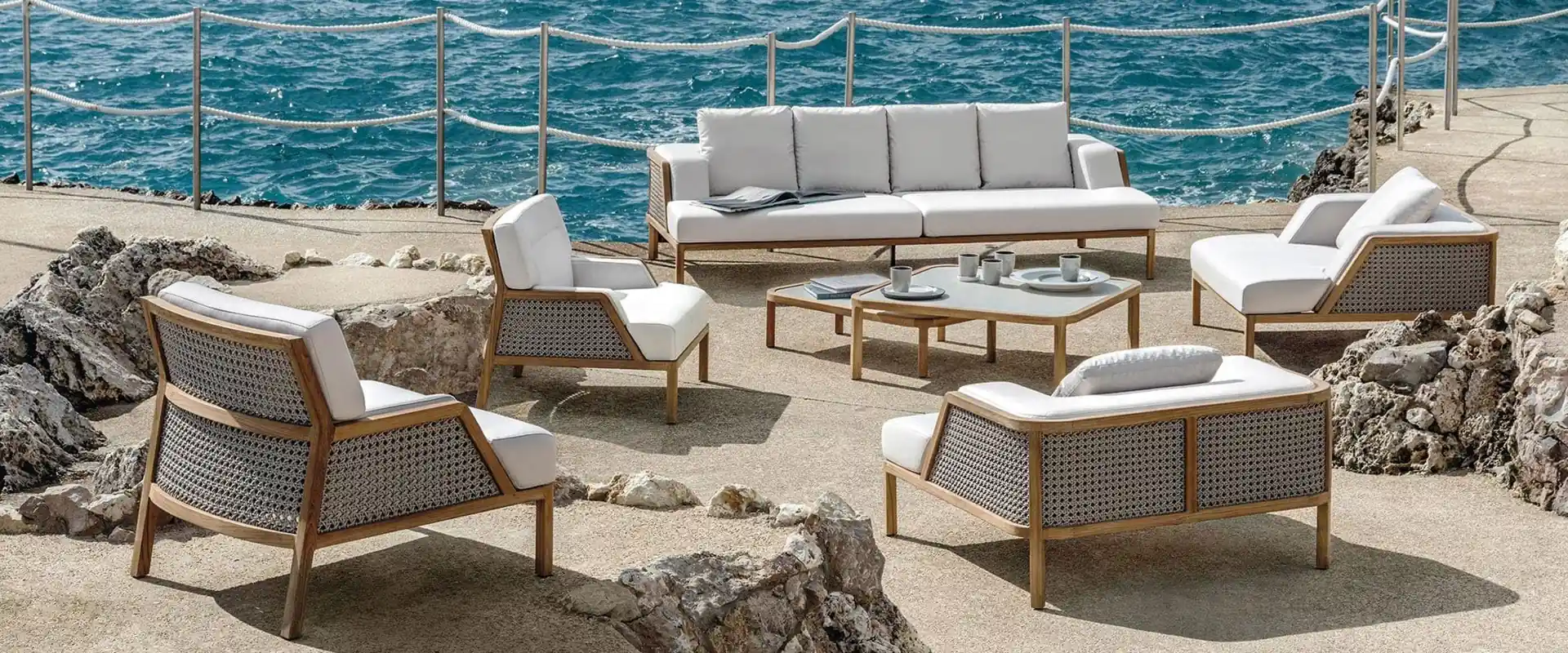 Ethimo Grand Life teak sofa and armchairs with white cushions around coffee tables on a rocky platform beside the sea