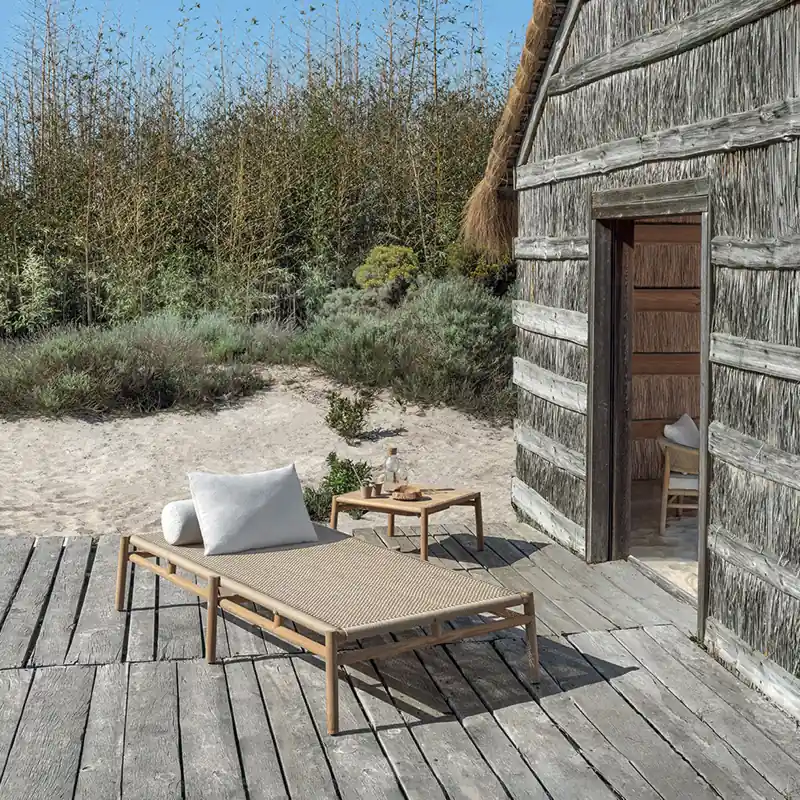 Ethimo Kilt teak daybed and side table on a weathered deck beside a wooden cabin among reeds