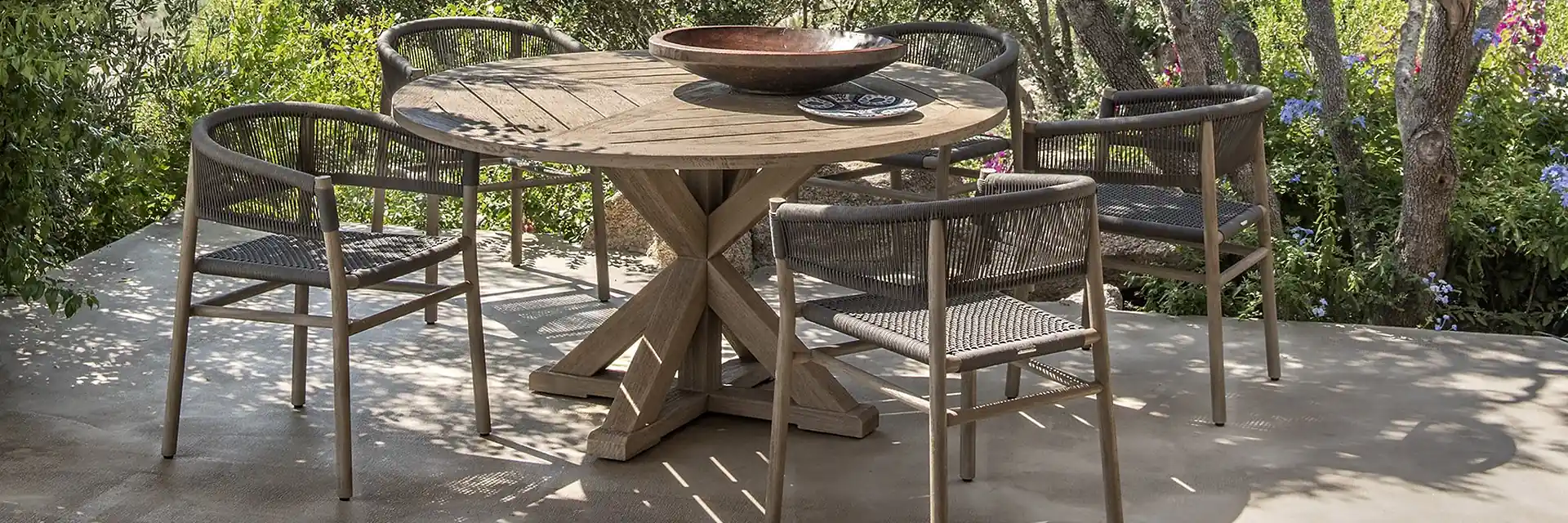 Ethimo Kilt rope and teak dining armchairs around a round teak table on a terrace shaded by trees, with a rocky hillside behind