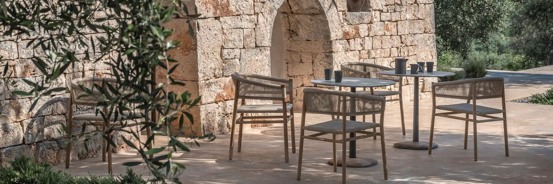 Ethimo Kilt rope and teak dining armchairs around round tables on a terrace beside a stone trullo, among olive trees