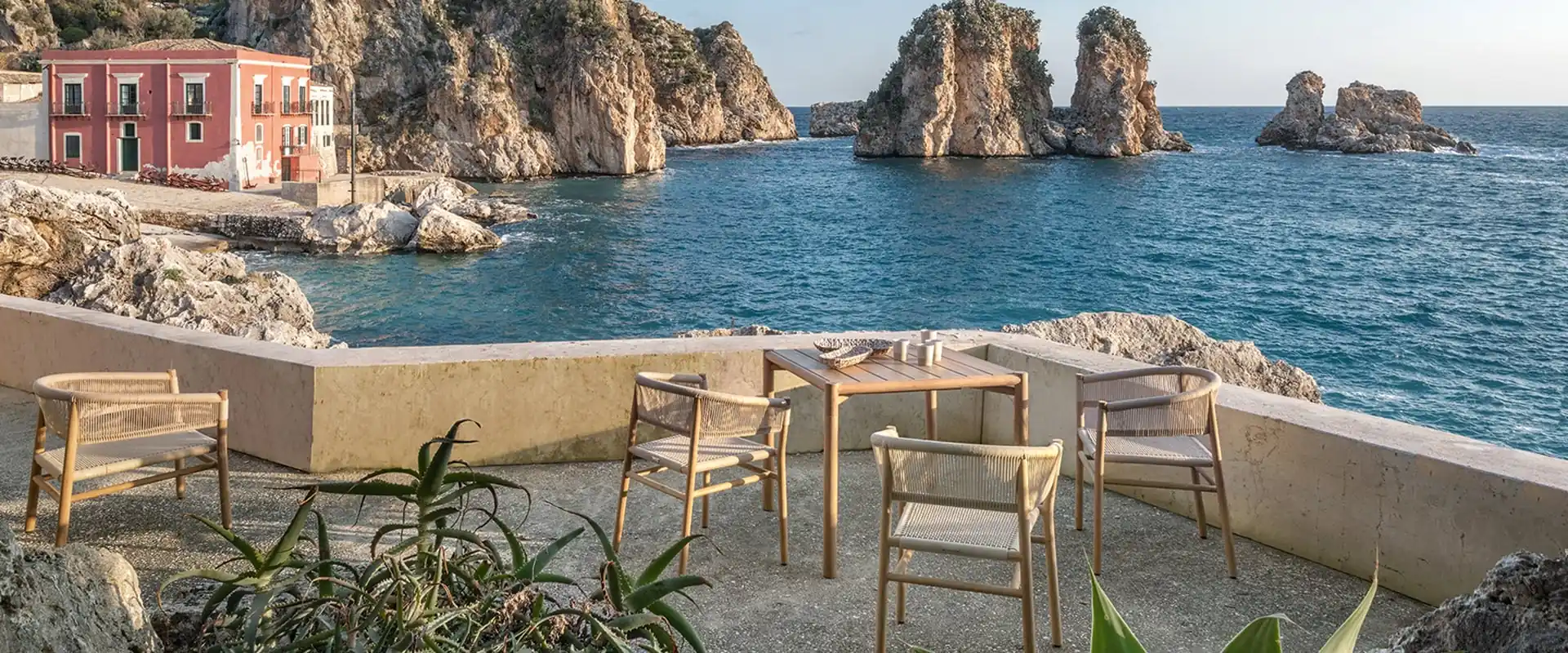 Ethimo Kilt teak dining table, chairs and bench on a stone terrace above a blue bay with sea stacks and a pink villa