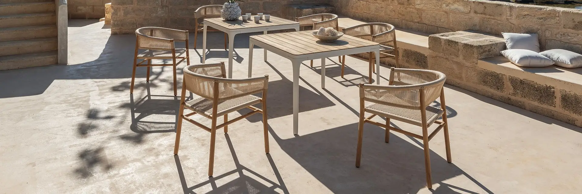 Ethimo Kilt teak dining tables and rope armchairs on a stone terrace overlooking the sea and a mountainous coastline
