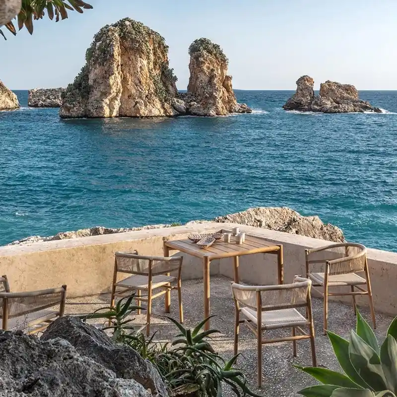 Ethimo Kilt teak dining table and chairs on a stone terrace, with sea stacks rising from a blue bay