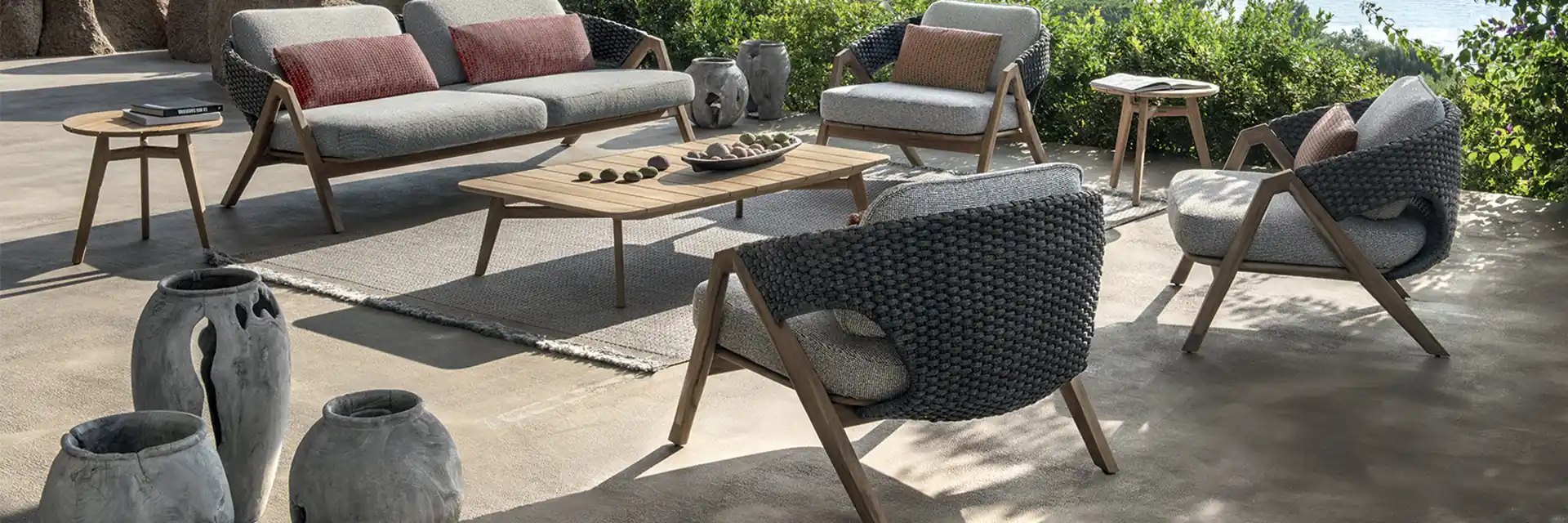 Ethimo Knit teak sofa and rope armchairs with red cushions on a terrace among granite rocks, above the sea