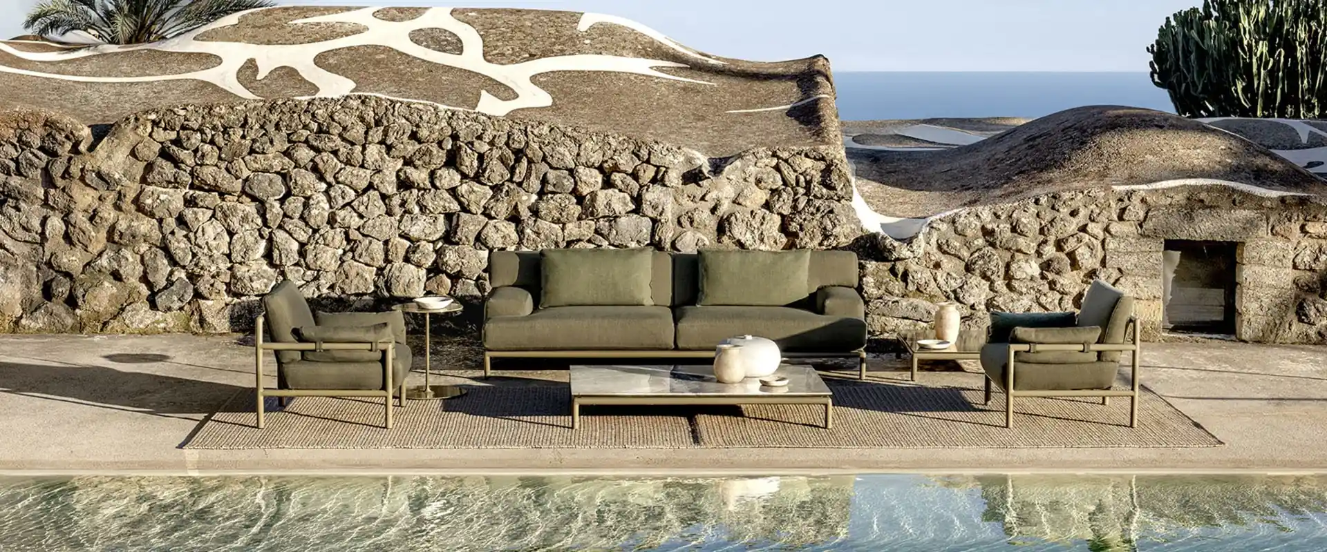 Ethimo Lodge sofa and armchairs with olive-green cushions before a dry lava-stone wall, with a pool in the foreground