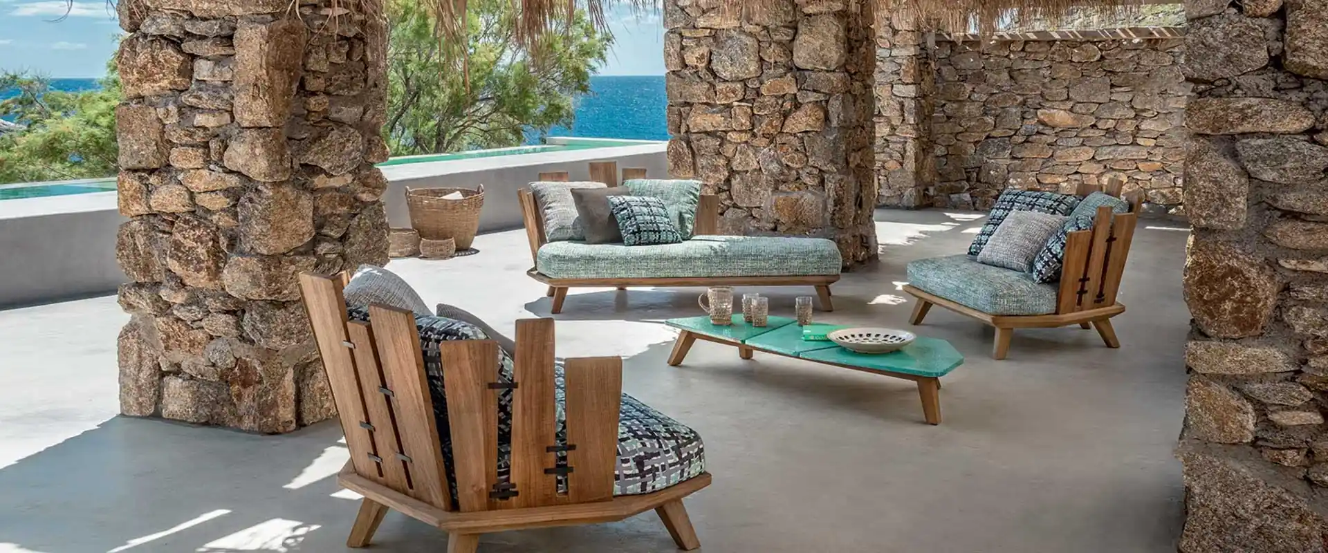 Ethimo Rafael teak daybed and lounge armchairs with patterned cushions under a stone and thatch pergola overlooking the sea
