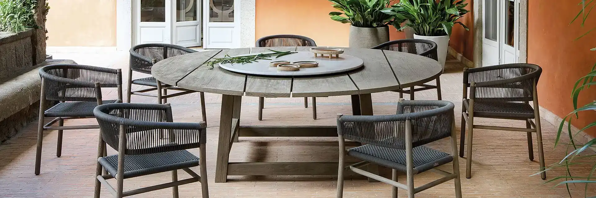 Ethimo Rafael round teak dining table with a lazy Susan, surrounded by rope chairs on the patio of a terracotta-coloured house