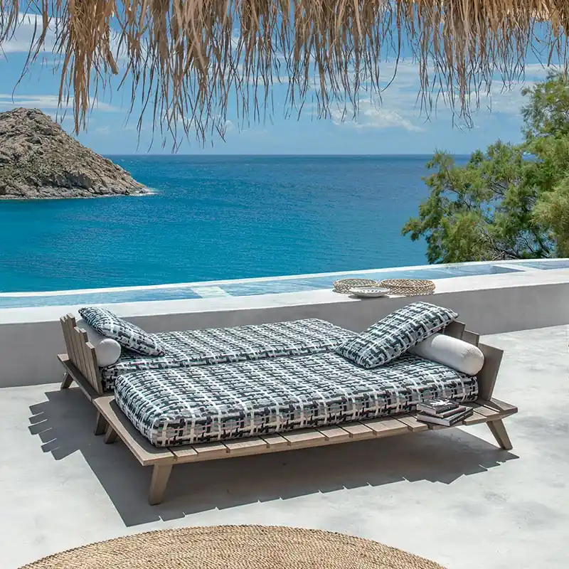 Two Ethimo Rafael teak sunbeds with checked mattresses on a terrace under a thatched roof, above a blue sea