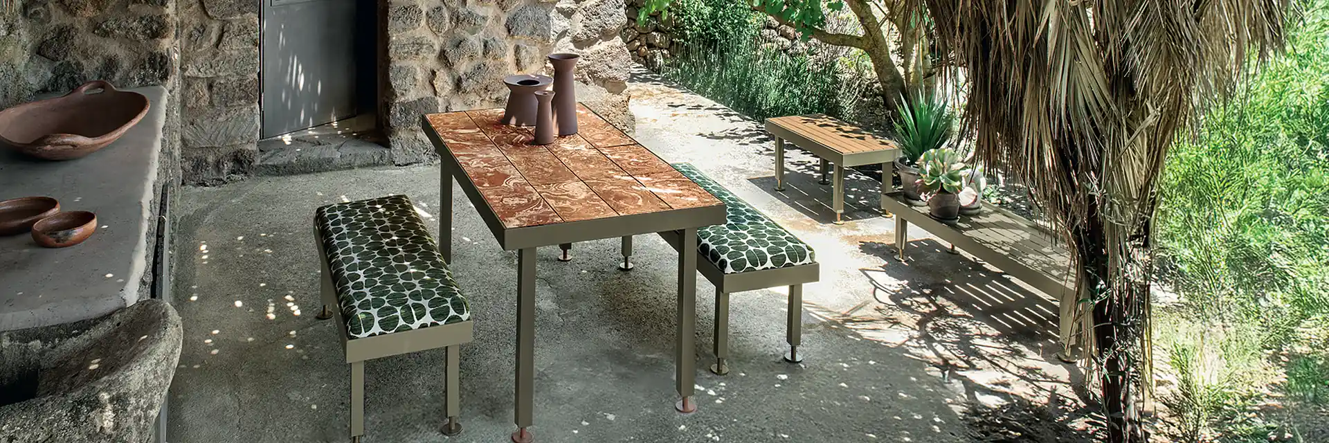 Ethimo Seeds table and benches with leaf-print cushions under a palm-thatched porch beside a stone house