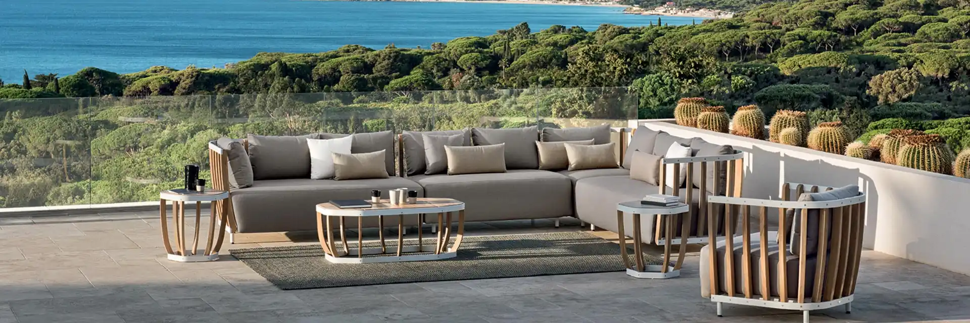 Ethimo Swing modular sofa and teak-slatted armchair on a terrace with cacti, overlooking a wooded bay