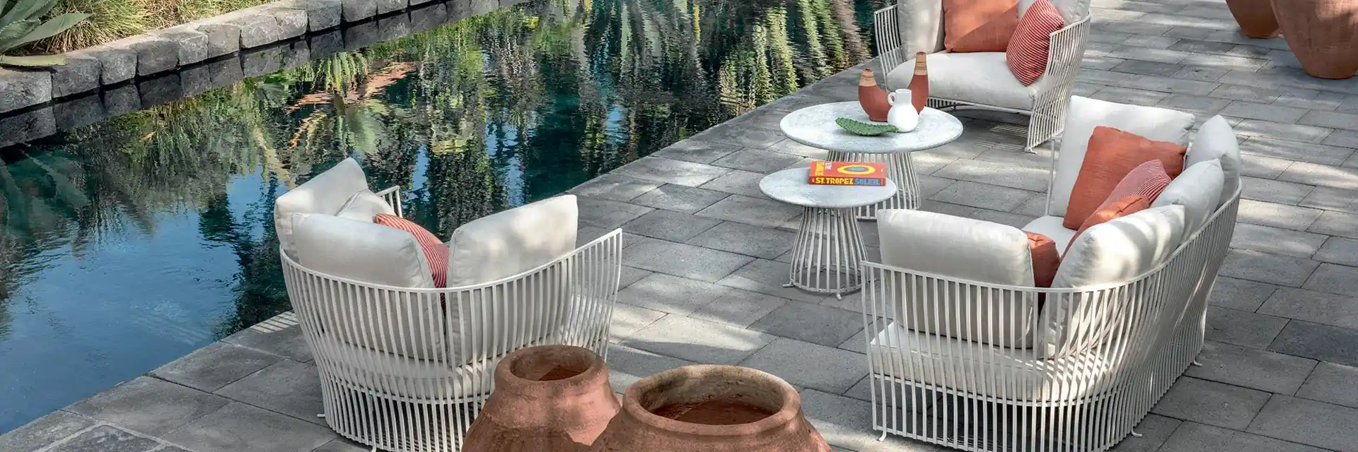 Ethimo Venexia white armchairs with coral cushions and round tables beside a dark stone pool, with terracotta jars