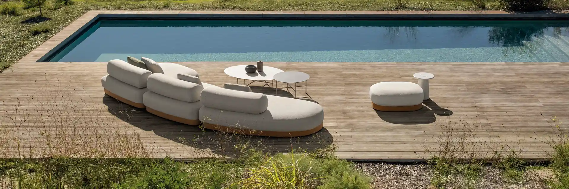 Expormim Brae curved modular sofa and ottoman on a wooden deck beside a long pool, with meadows and trees beyond