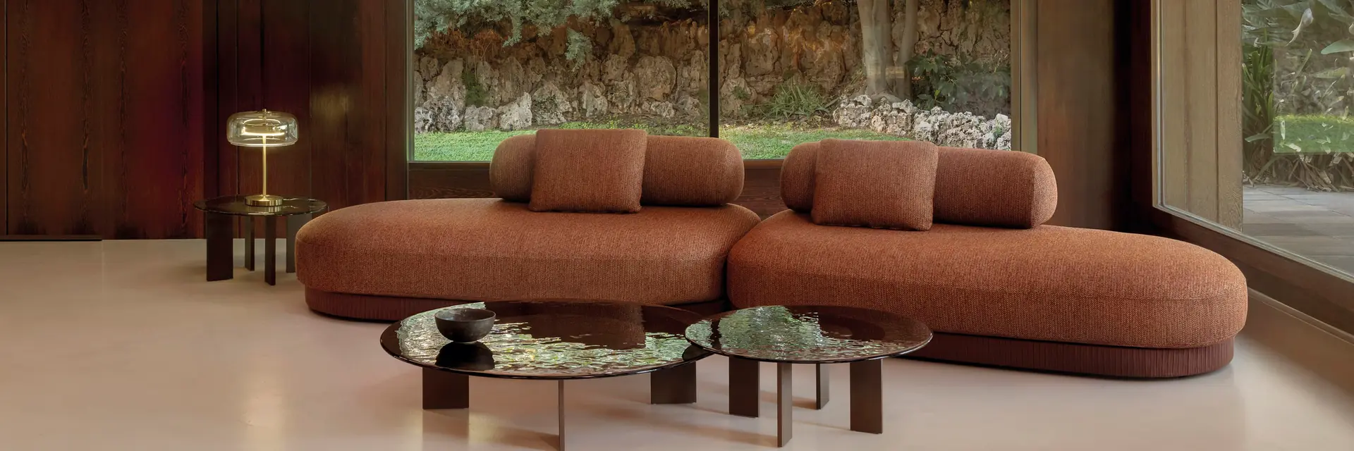 Expormim Brae curved sofa in terracotta fabric with round glass coffee tables, in a timber-panelled room with a wide garden window