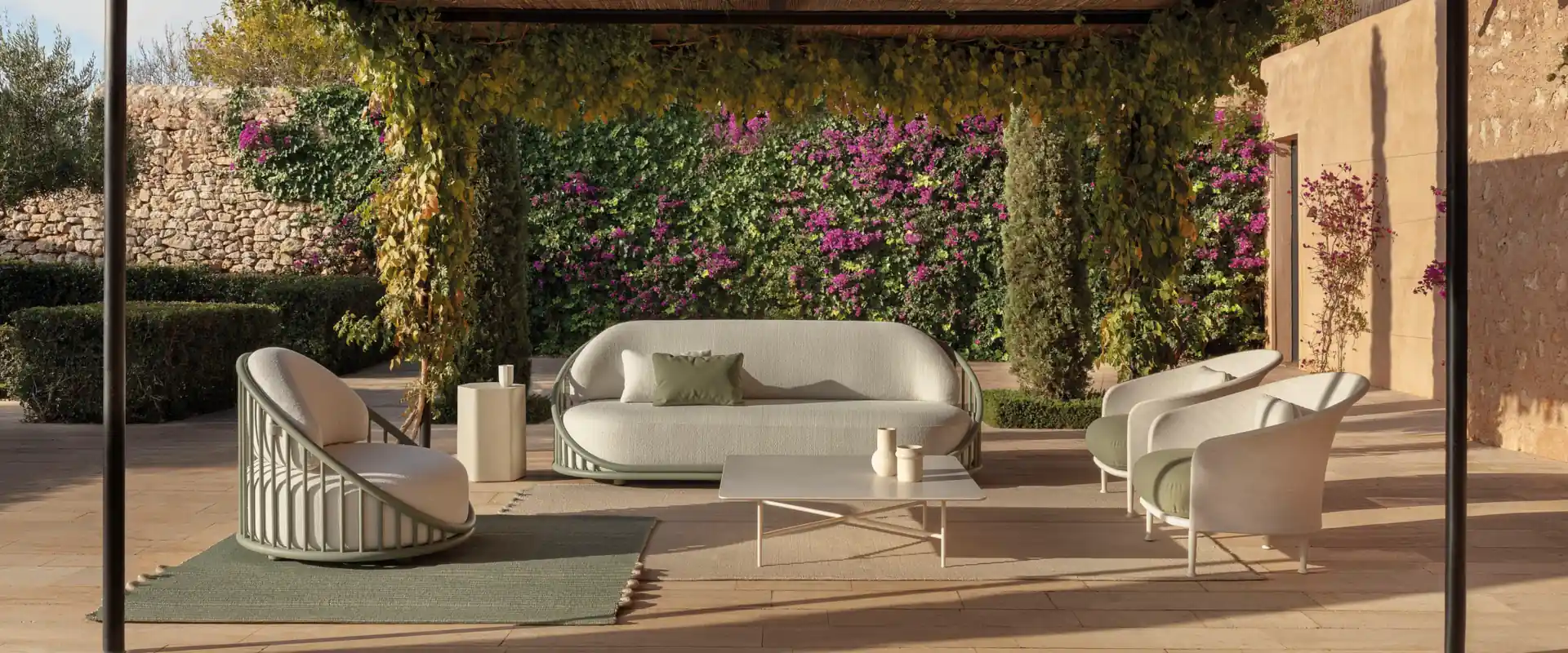 Expormim Cask sofa and armchairs under a dark pergola, with a wall of ivy and pink bougainvillea behind