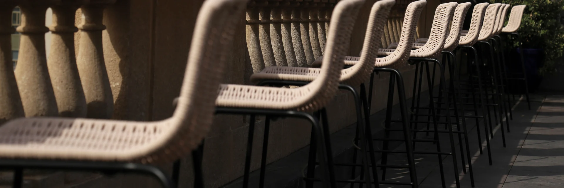 Row of Expormim Lapala woven bar stools along a stone balustrade on a rooftop terrace, in low sun