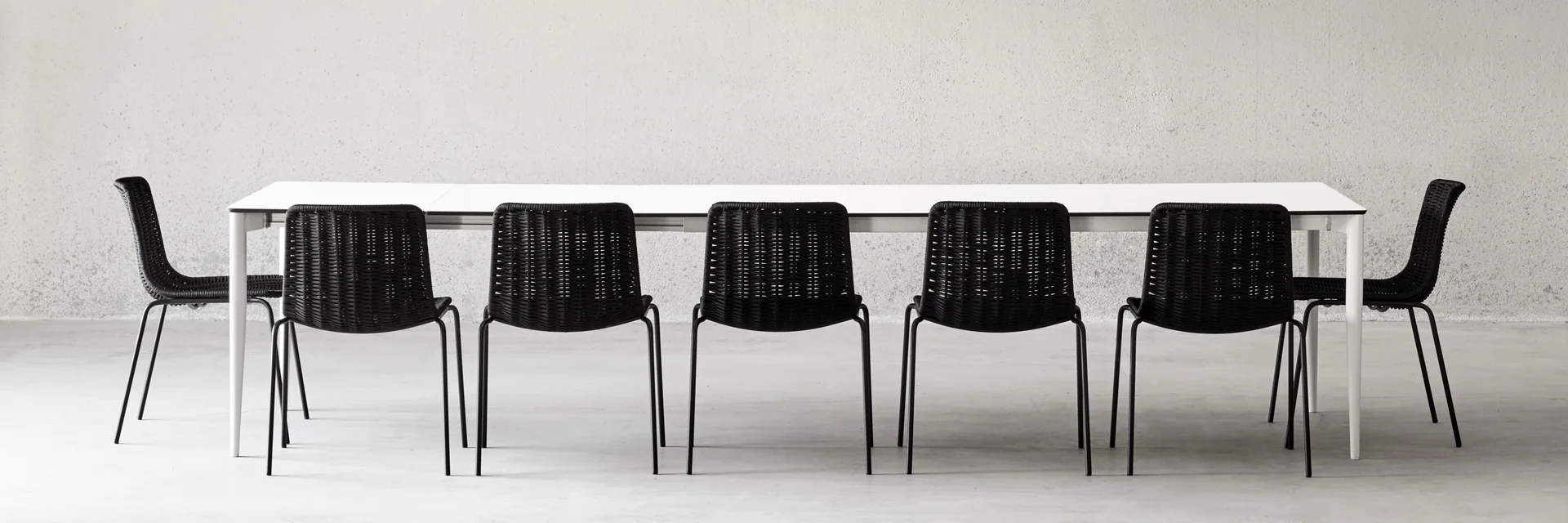 Black Expormim Lapala woven chairs along a long white table, against a pale concrete wall