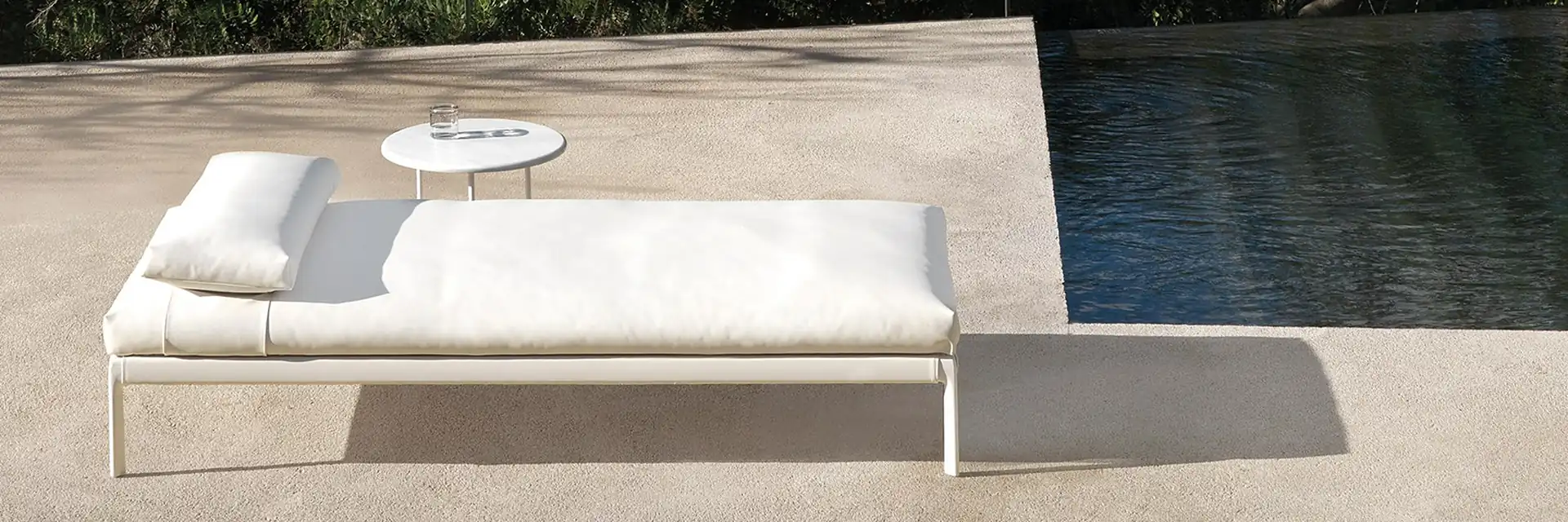 White Expormim Livit daybed with a small side table at the edge of a dark pool, in the shade of trees