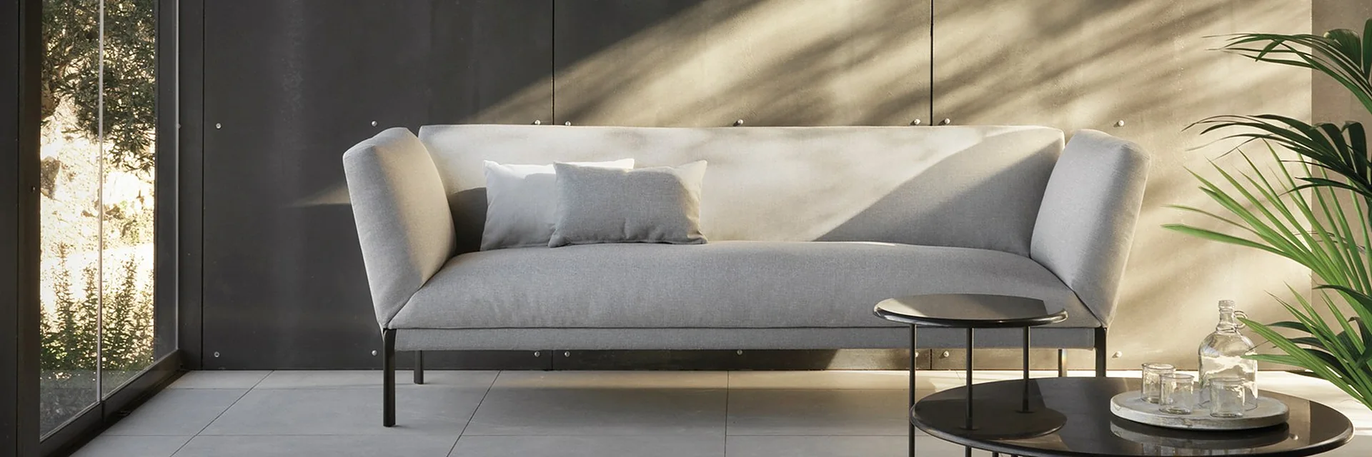 Expormim Livit sofa with pale cushions and round low tables on a terrace beside a dark wall, in dappled shade