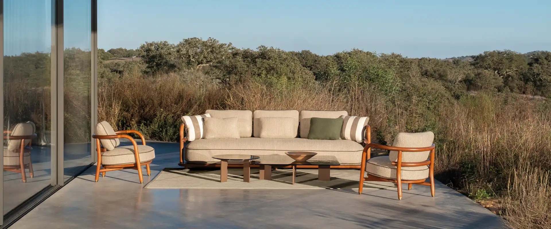 Expormim Meridies sofa and wooden-framed armchairs with striped cushions on a stone terrace under a flat roof, overlooking oak-covered hills