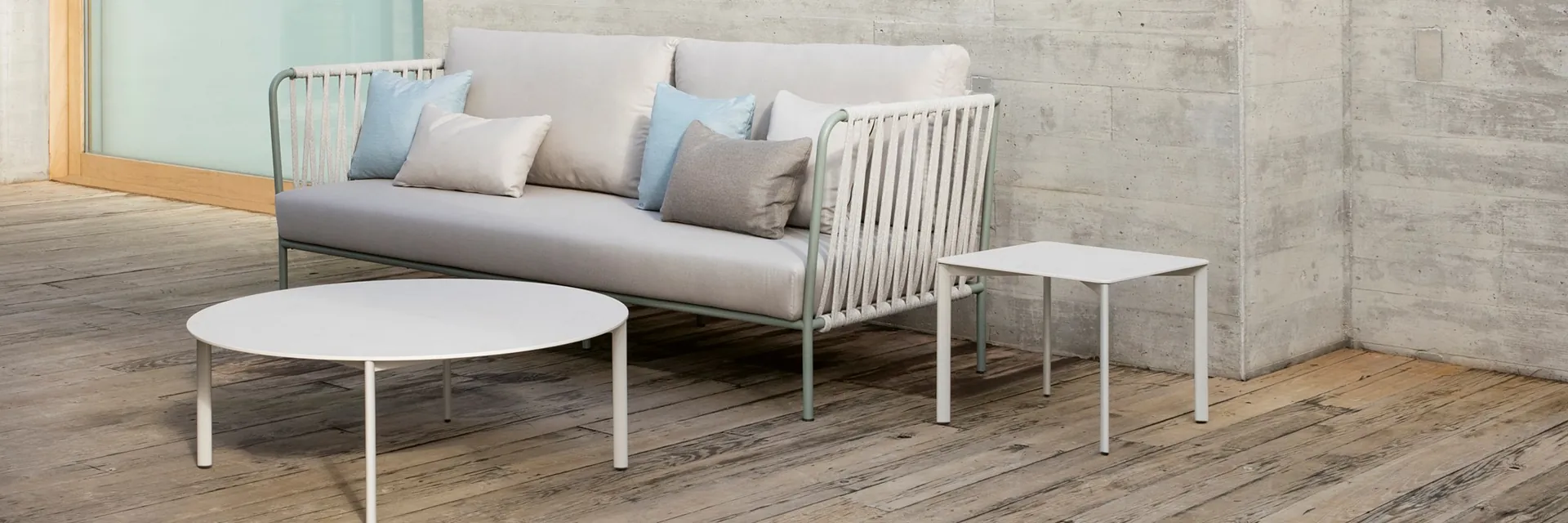 Expormim Nido sofa with a sage-green frame and white cord back, beside two low tables on a weathered timber deck by a concrete wall