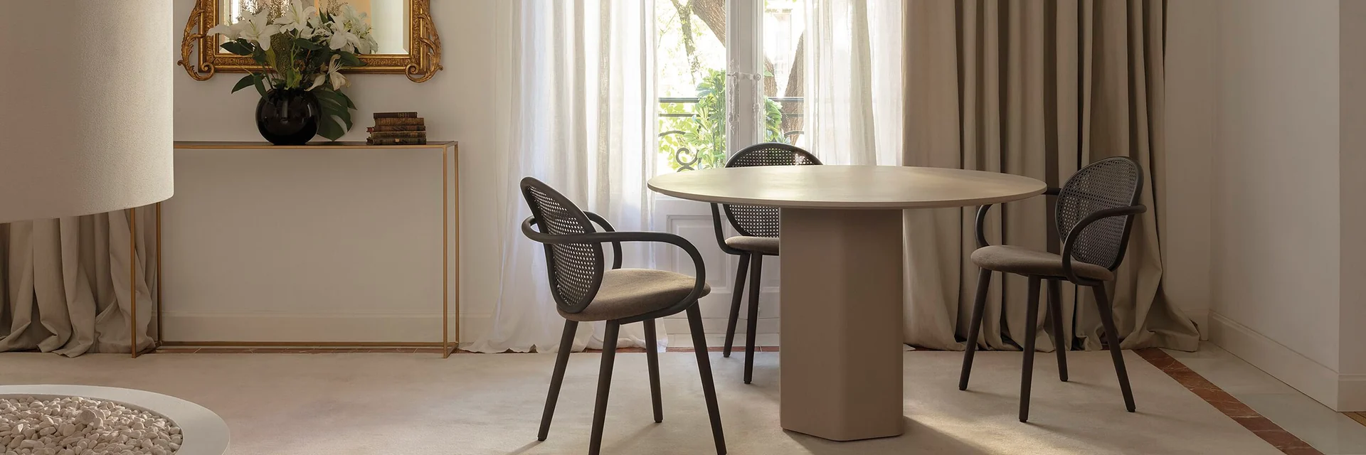 Expormim Talo round table with dark cane-backed chairs in a bright room with tall windows, linen curtains and a gilt mirror