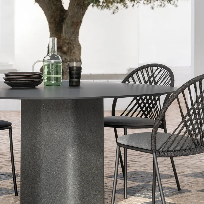 Expormim Talo round dining table with a dark fluted base and black woven chairs on a patio under an olive tree