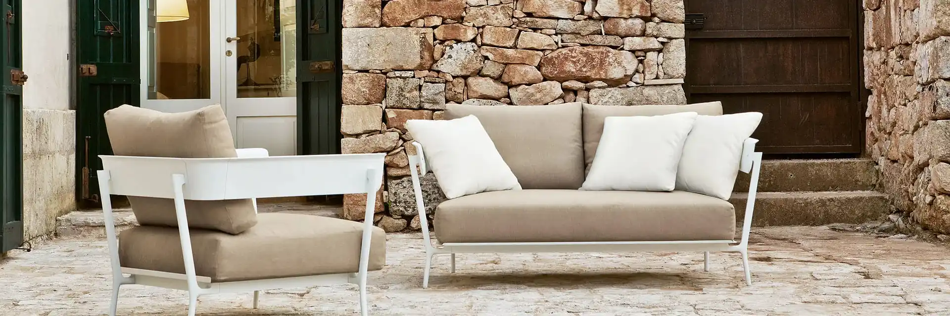 Fast Aikana armchair and two-seater sofa with white frames and taupe cushions in a courtyard with a dry-stone wall