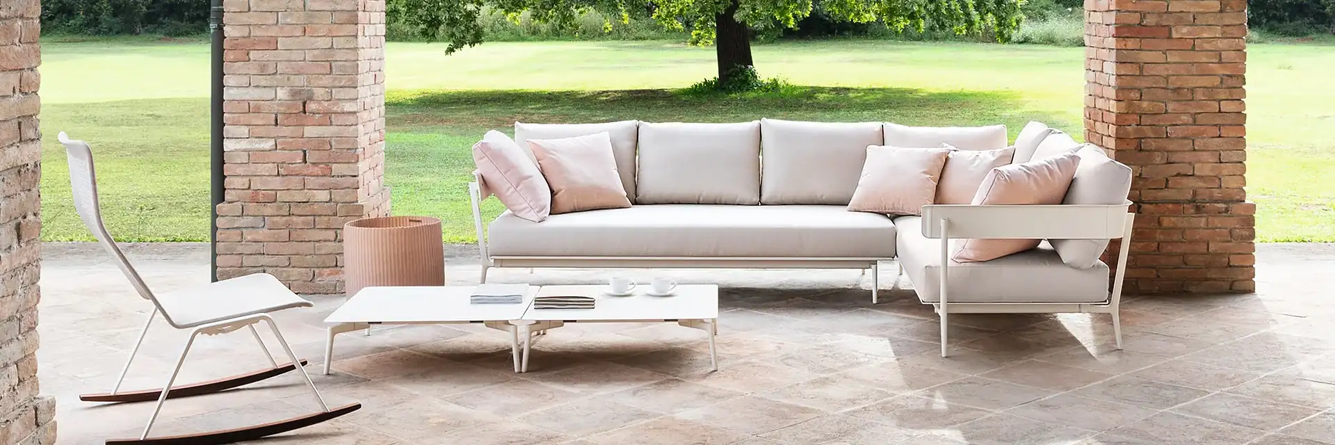 Fast Aikana corner sofa with pale cushions and low tables under a brick portico, with a lawn and a large tree behind