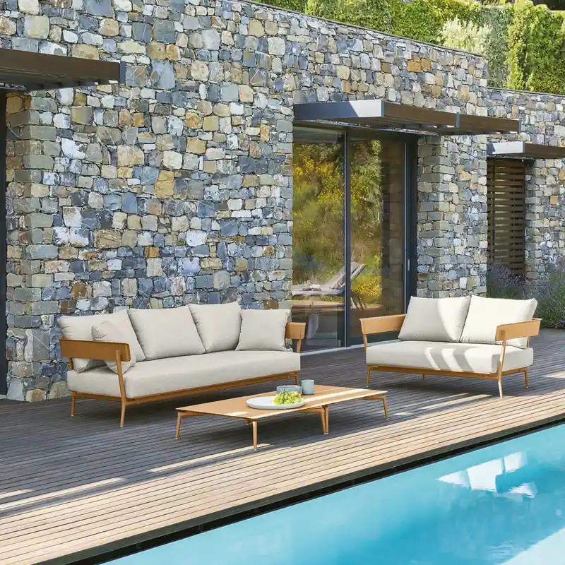 Fast Aikana aluminum sofas with cream cushions on a wooden deck beside a pool, in front of a stone house covered in greenery