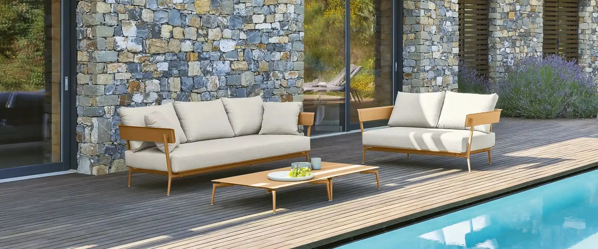Fast Aikana aluminum sofas with cream cushions on a wooden deck beside a pool, in front of a stone house covered in greenery