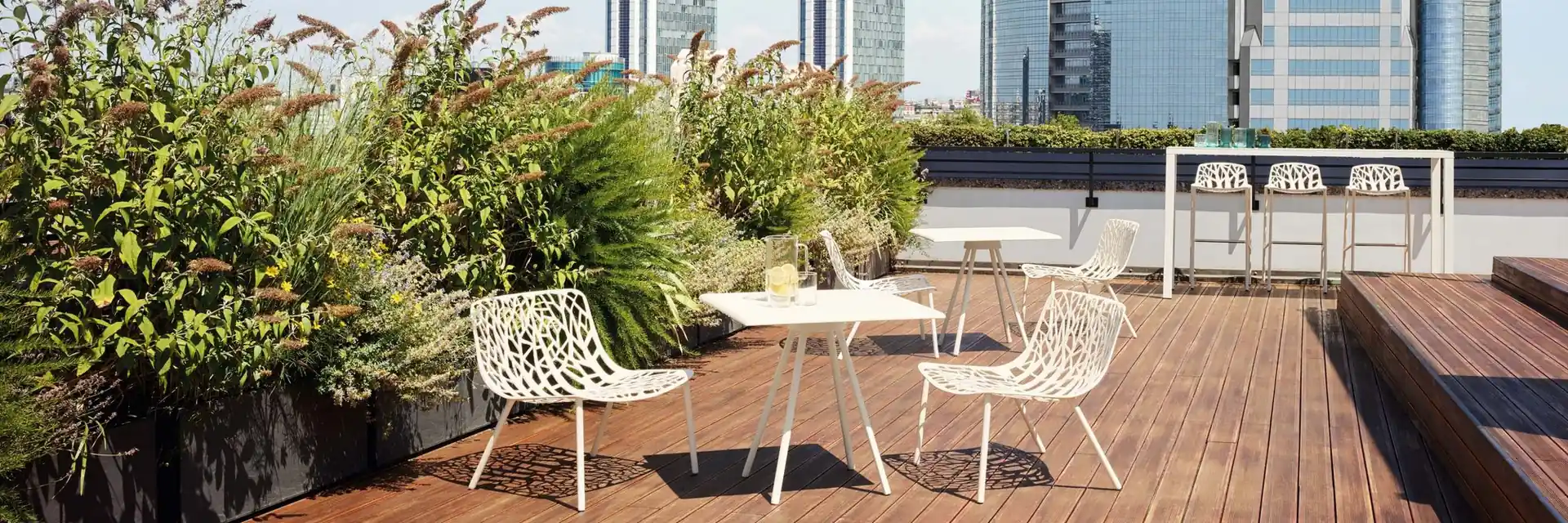 White Fast Forest armchairs and bar stools on the timber deck of a planted rooftop terrace, with city towers behind