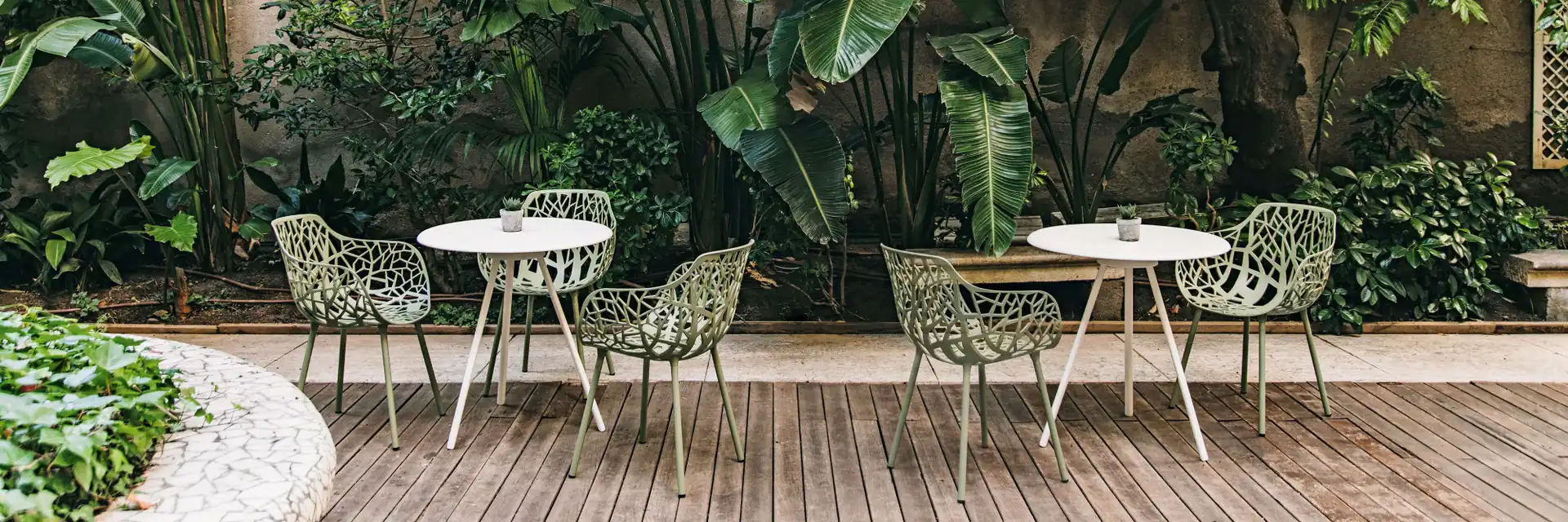 Fast Forest aluminium armchairs in pale green around two round tables on a timber deck, in a courtyard full of tropical plants
