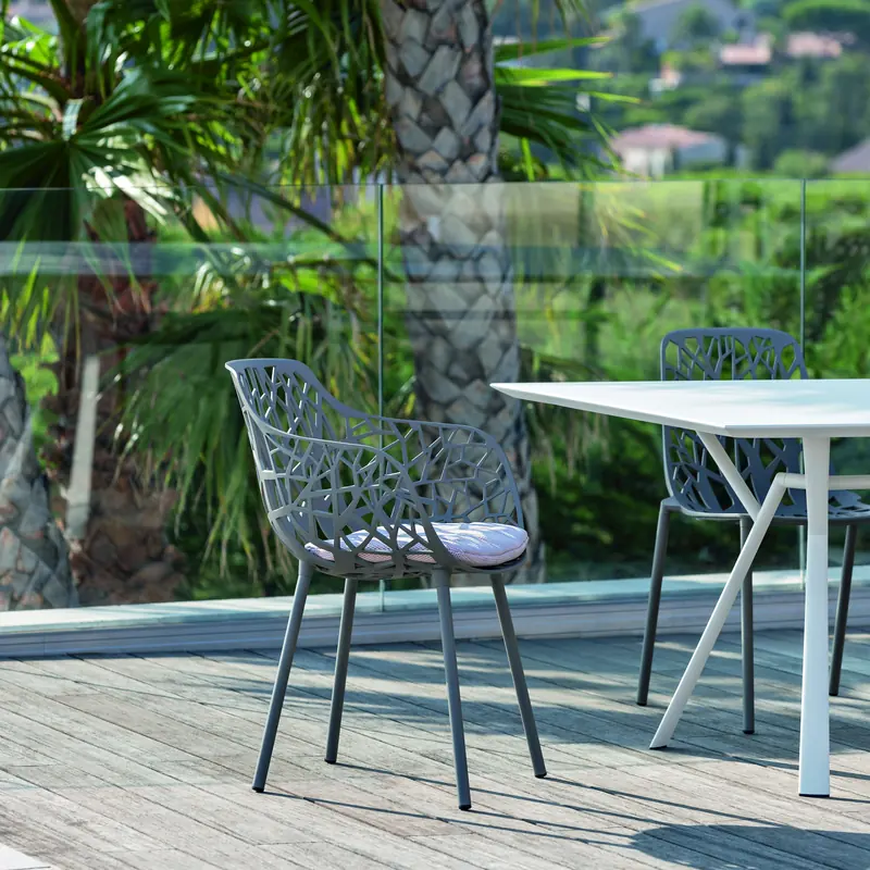 Fast Forest aluminum chair with a branch-pattern back at a white table on a wooden deck, palm trees and hills behind