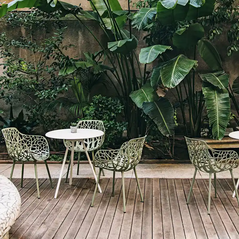 Fast Forest white aluminum chairs with branch-pattern backs at round tables on a wooden deck, in front of lush tropical plants