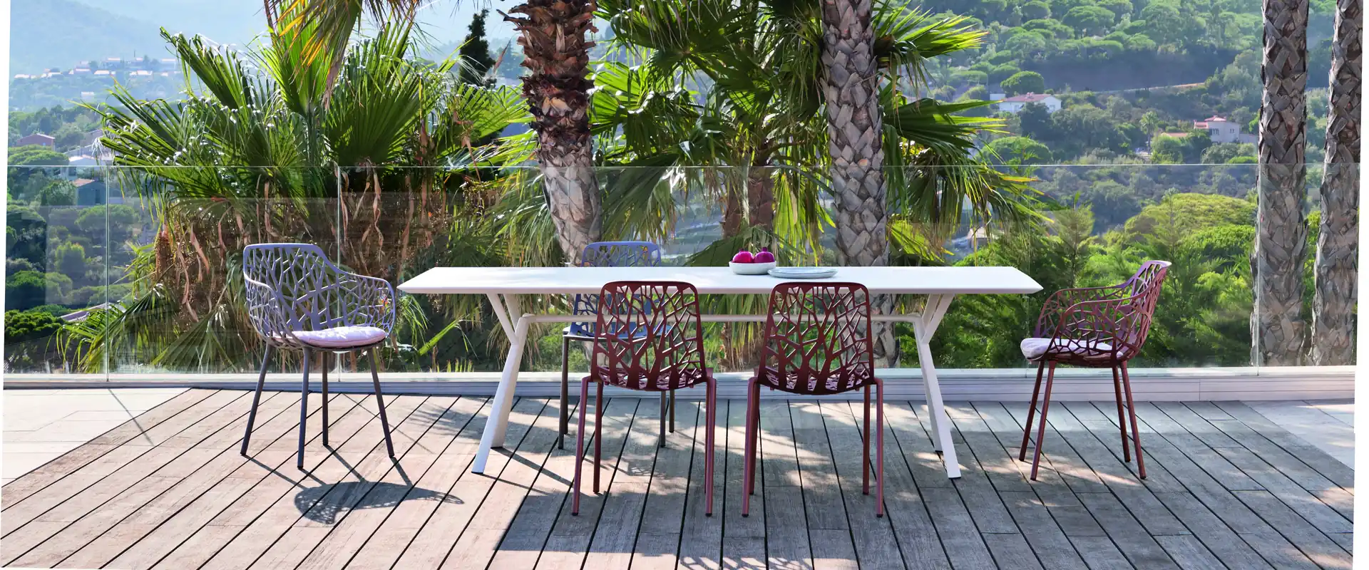Fast Forest aluminum chairs with branch-pattern backs in burgundy around a long white Radice Quadra table on a wooden deck, with palm trees and hills behind