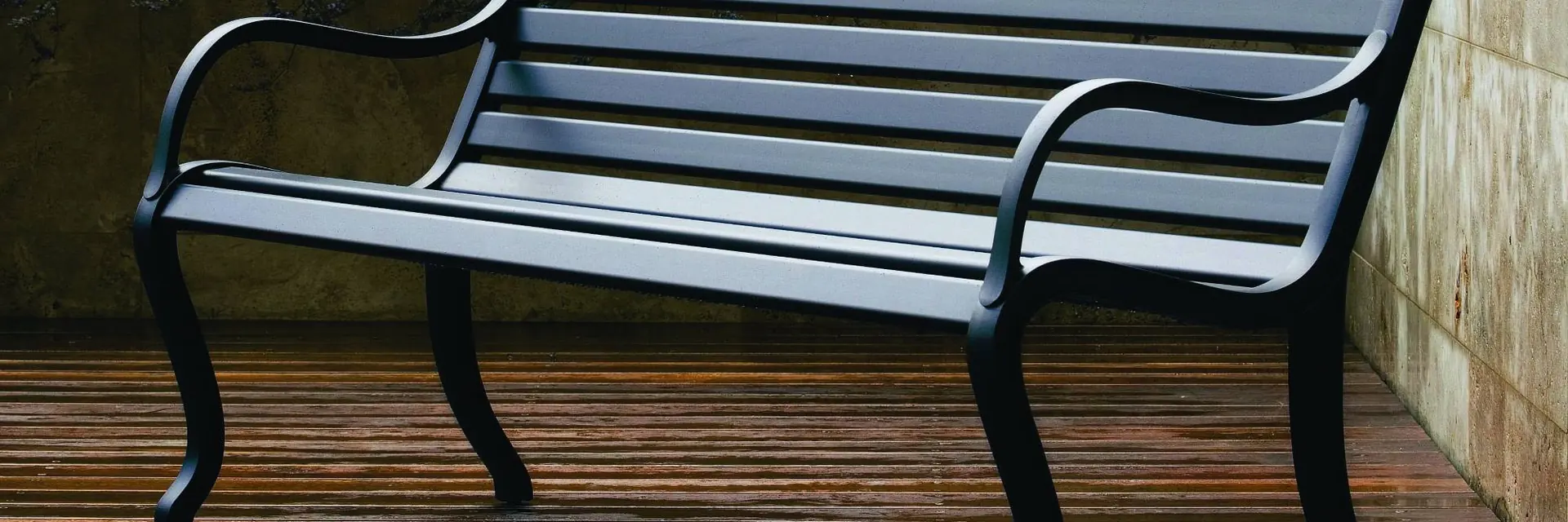 Fast Oasi dark aluminum bench on a wooden deck beside tall grasses and a stone wall
