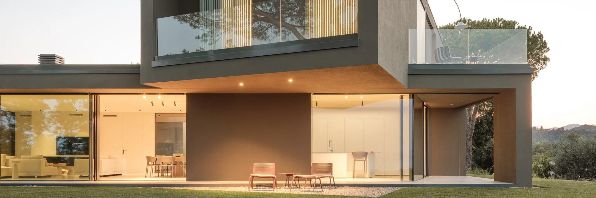 Fast outdoor chairs on the upper terrace of a modern house with a lit interior, seen from the lawn at dusk