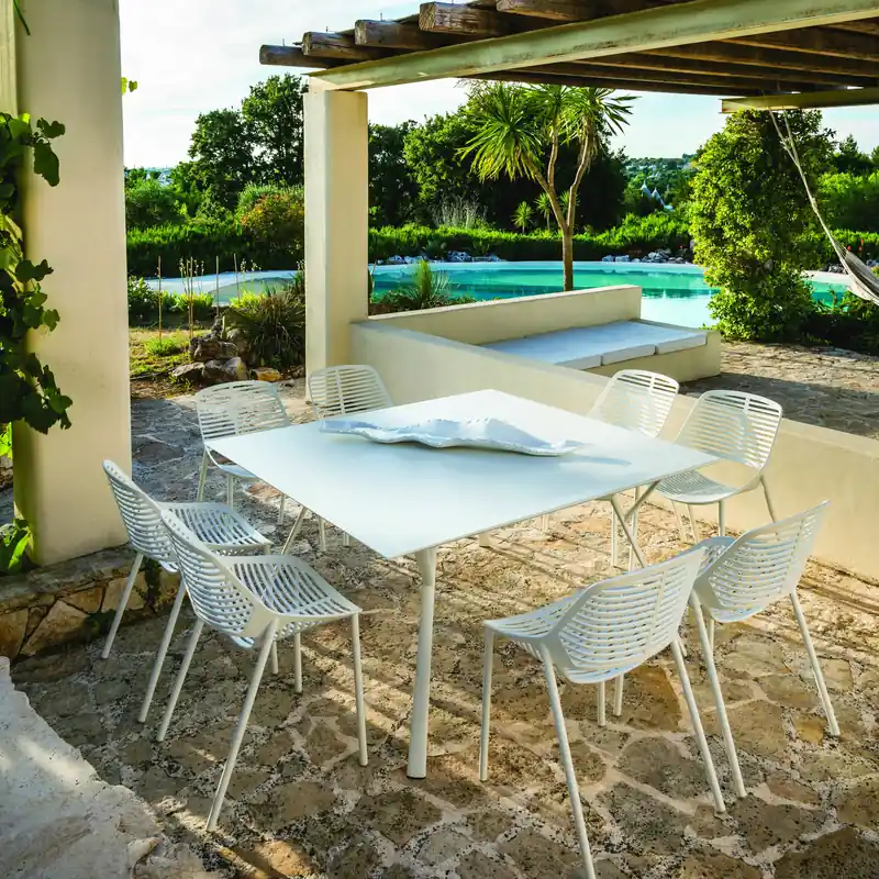 Fast Radice Quadra square white table with white slatted chairs under a wooden pergola, with a pool and palm trees behind