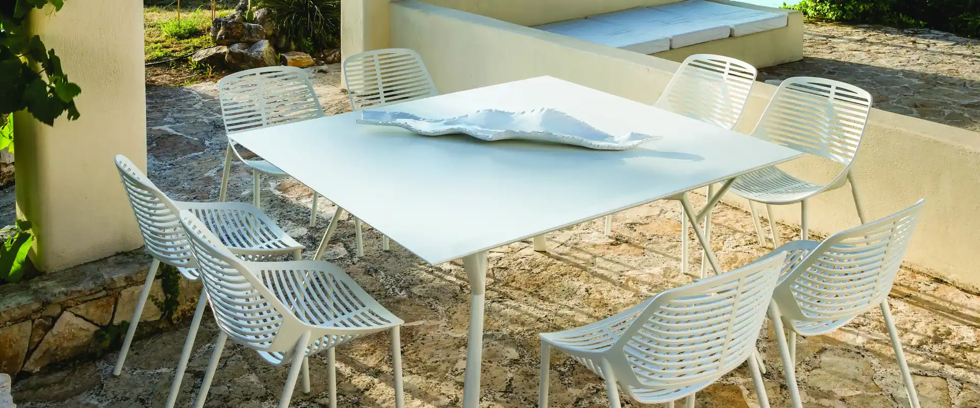 Fast Radice Quadra square white table with white slatted chairs under a wooden pergola, with a pool and palm trees behind
