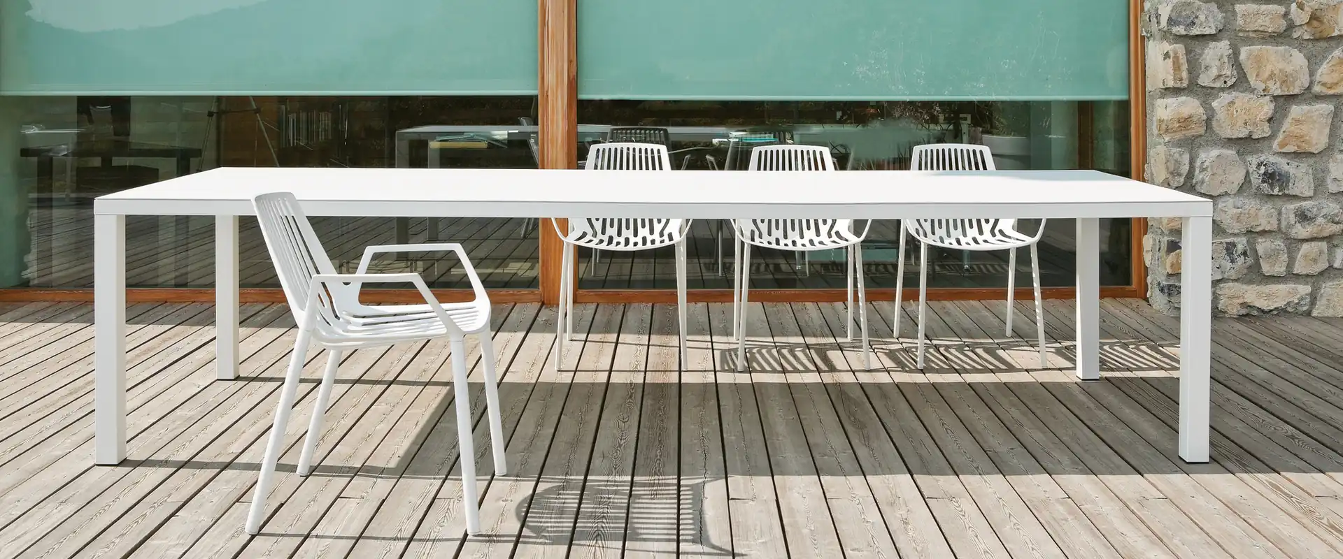 White Fast Rion aluminum chairs around a long white dining table on a timber deck, in front of a glazed wall and a stone house