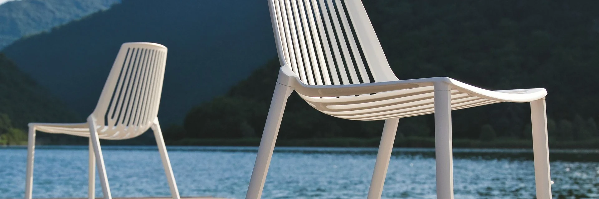 Two white Fast Rion aluminium chairs on a timber deck beside a lake, with wooded mountains behind