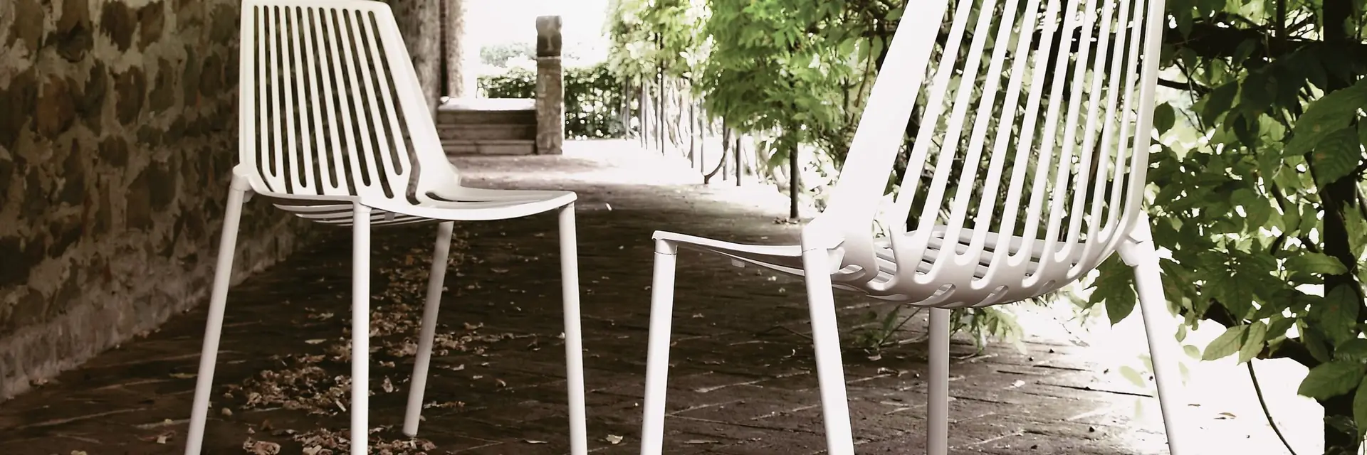 White Fast Rion aluminium chairs on a paved path beside a stone wall, under the leaves of a fig tree