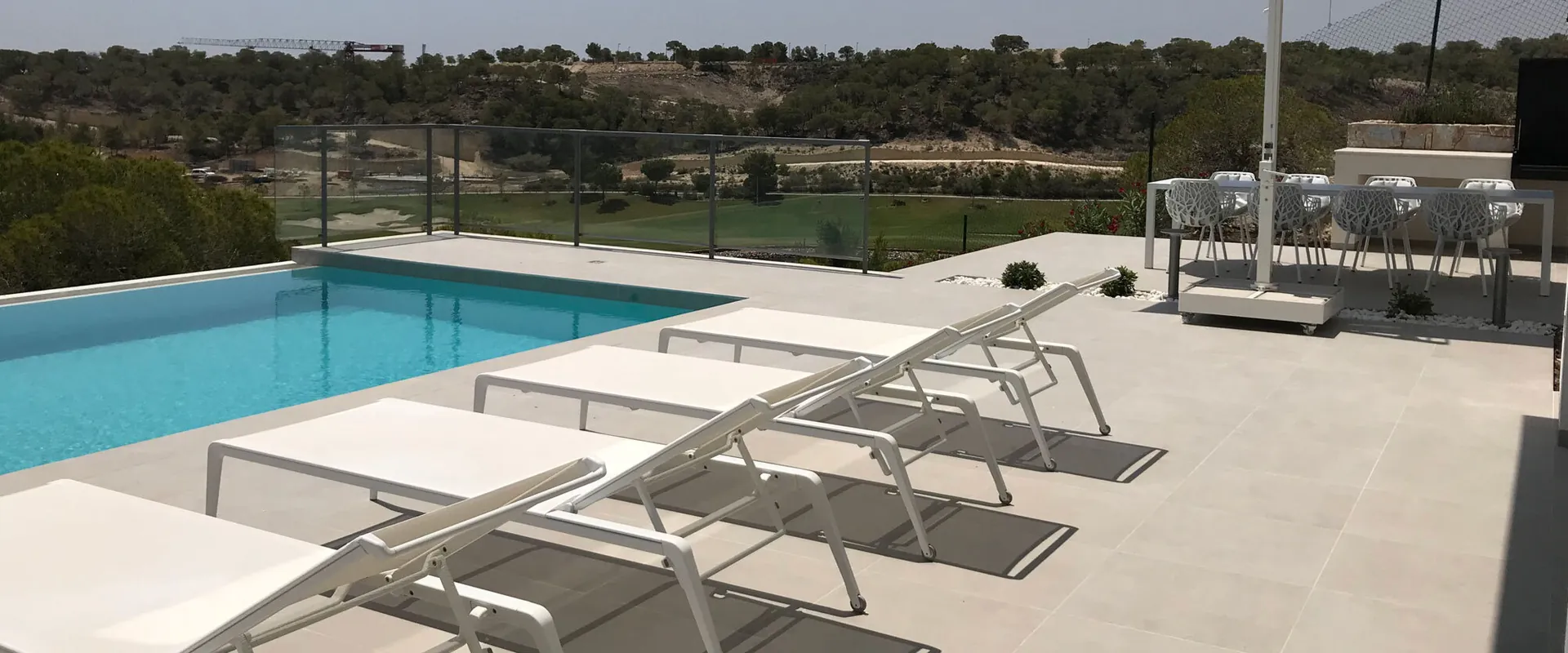 Fast aluminum sun loungers with white slings lined up beside a pool, with a parasol and scrub-covered hills behind
