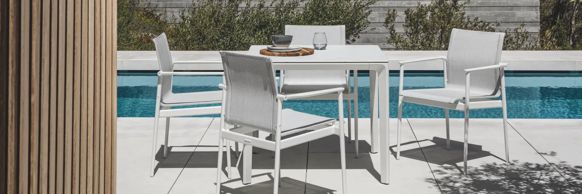 White Gloster 180 stacking chairs around a dining table on a terrace beside a pool, in front of a board-marked concrete wall