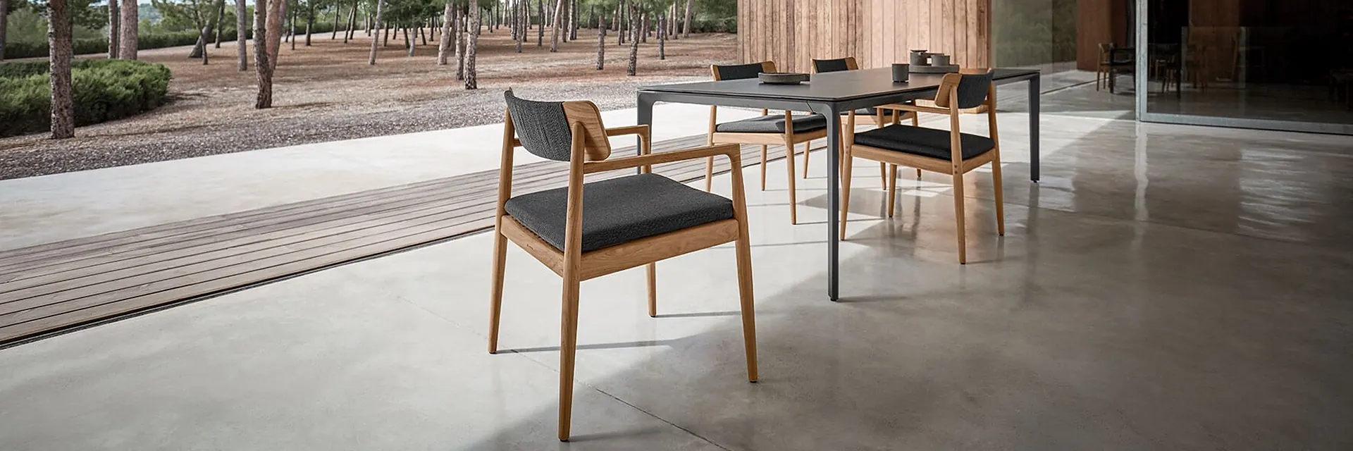Gloster Archi dining chairs and table on a polished concrete terrace under a timber roof, with pines beyond