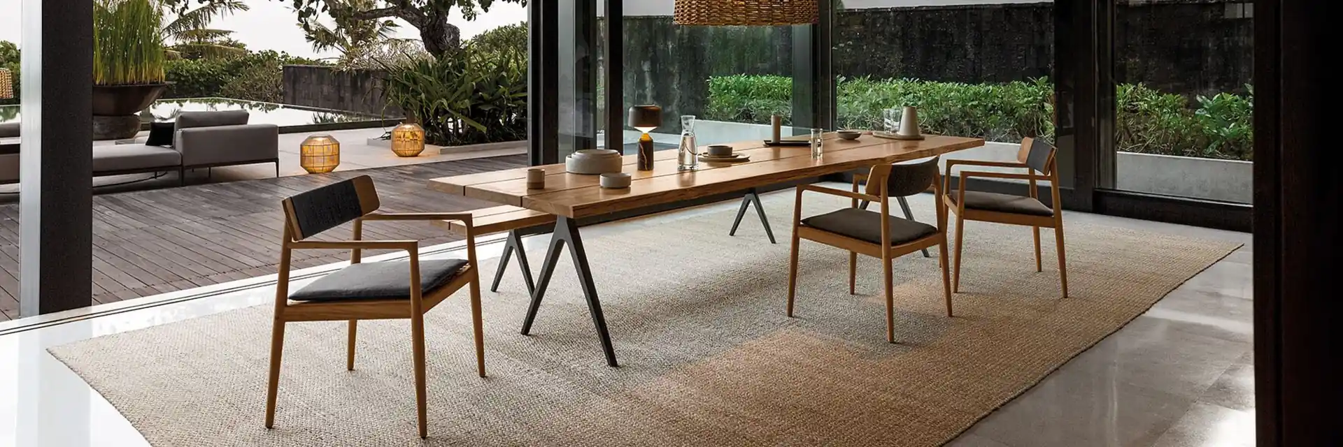 Gloster Archi teak dining table and chairs on a rug under a dark steel pergola, with a garden beyond