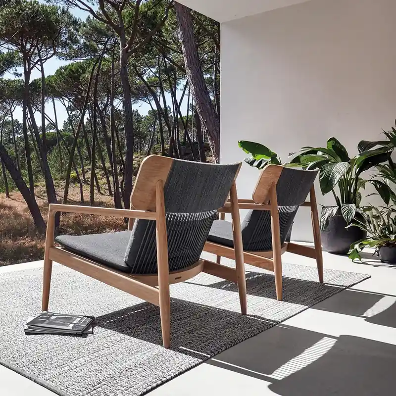 Gloster Archi teak lounge chairs with dark woven seats on a rug, on a terrace opening onto a pine wood